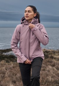 Young woman wearing a mauve jacket and black pants zips up outdoors near a rocky shoreline under a cloudy sky.