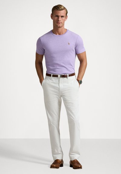 Λιλά κοντομάνικο μπλουζάκι με στρογγυλή λαιμόκοψη, συνδυασμένο με λευκά παντελόνια και καφέ δερμάτινα παπούτσια. Διαθέτει ένα μικρό λογότυπο στο στήθος.