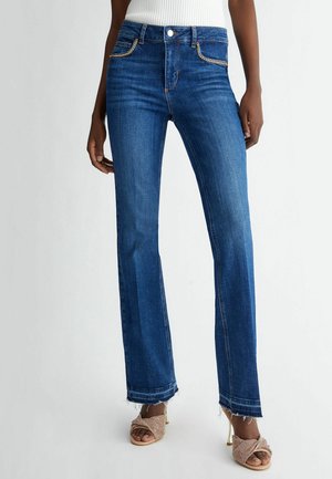 Femme portant un jean bleu droit avec des ourlets effilochés, un haut blanc côtelé et des sandales à talons hauts ouvertes beige sur un fond uni.