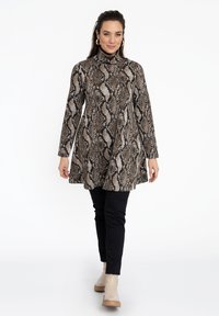 Bruine tuniek met slangenpatroon, hoge kraag, lange mouwen en uitlopende zoom; gecombineerd met zwarte strakke broek en beige enkellaarzen.