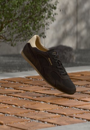 Donkerbruine suède sneaker met beige hiel en zwarte veters, zwevend boven een houten tegelvloer met grijze kiezels buiten.