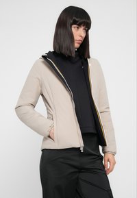 Giacca imbottita beige con zip frontale, polsini elasticizzati e una fodera interna colorata, indossata sopra un maglione nero e pantaloni neri.
