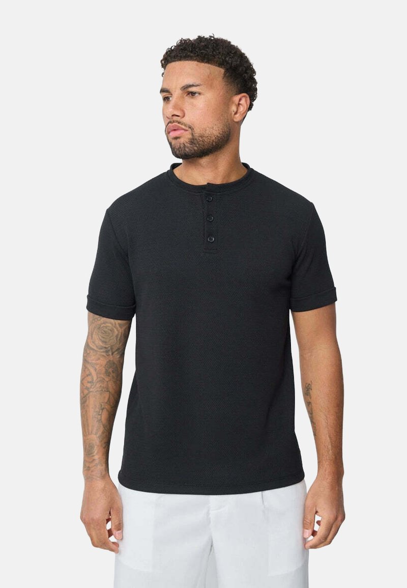 Chemise henley noire à manches courtes avec patte de boutonnage à trois boutons, tissu texturé et manches à revers, portée par un homme tatoué.