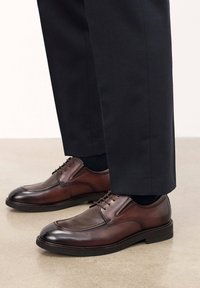 Zapatos de piel marrón con cordones, acabado suave y paneles texturizados, combinados con pantalones oscuros y calcetines negros sobre una superficie neutra.