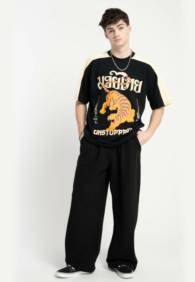 Młody mężczyzna stojący z rękami w kieszeniach, ubrany w czarne spodnie, czarno-beżową koszulę z wzorem pomarańczowego tygrysa i napisem "NIE DO ZATRZYMANIA"