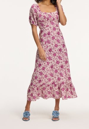 Kvinde iført en lyserød blomstrede midi-kjole med pufærmer og flæsekant, kombineret med blå åbne slip-on sandaler med blomsterudsmykninger.