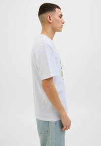 Hvid bomulds T-shirt med korte ærmer, der har et grafisk print i grøn og sort på fronten. Båret med lyseblå denimjeans.