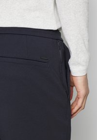 Marineblauwe stretchy broek met een platte achterzak met ritssluiting, een gladde textuur en een elastische tailleband, gecombineerd met een grijze langemouwen shirt.