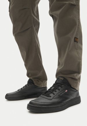 Jalad, mis on seljas oliivirohelised püksid ja mustad Reebok Classic tossud, millel on paelad, nähtavad põlve keskelt allpool valgel taustal.