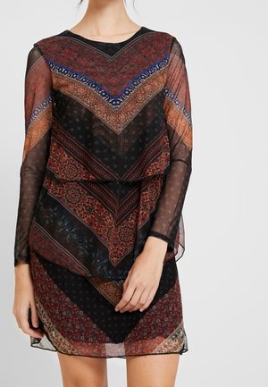 Femme portant une robe longue à manches longues en tissu transparent avec des motifs floraux géométriques superposés en noir, rouge, bleu et marron.