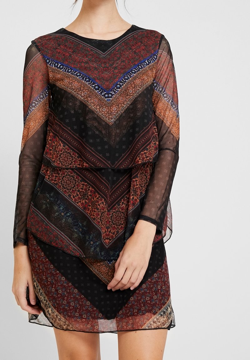 Femme portant une robe longue à manches longues en tissu transparent avec des motifs floraux géométriques superposés en noir, rouge, bleu et marron.