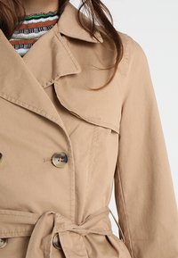 Trench-coat beige avec un design à double boutonnage, grand col et ceinture à nouer. Caractérisé par des boutons brillants et un tissu lisse et texturé.