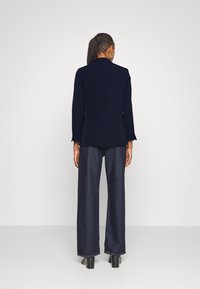 Blazer bleu marine avec une coupe ajustée, manches longues et une fente à l'arrière, associé à un pantalon en jean à jambes larges et des chaussures à talons.