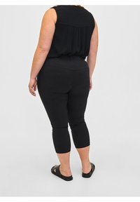 Zwarte stretchleggings met een aansluitend ontwerp, verkorte lengte en een gladde textuur, gecombineerd met een losse zwarte top. Eenvoudige schoenen maken de look compleet.