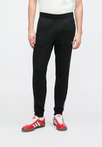 Čierne jogger nohavice s rebrovaným pásom, bočnými pruhmi a hladkým povrchom, skombinované s červenými atletickými topánkami s bielymi akcentmi.