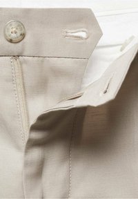 Gros plan sur la taille d’un pantalon beige avec bouton, boutonnière, fermeture éclair et rabat avant partiellement ouvert.
