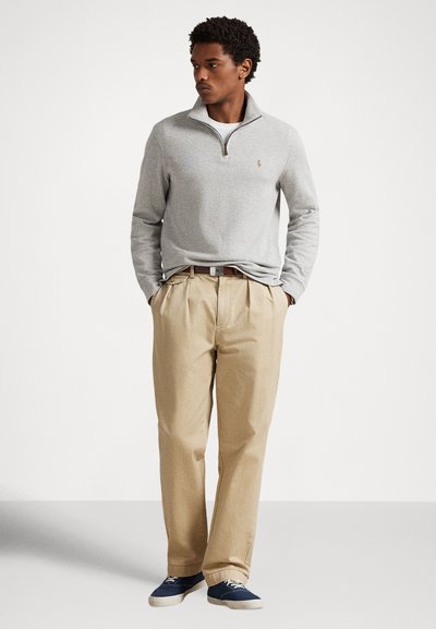 Sivi pulover z zadrgo na vratu, bež hlače z naborki, rjav pas in modre superge. Priložnostna obleka z mehko tkanino in sproščenim krojem.