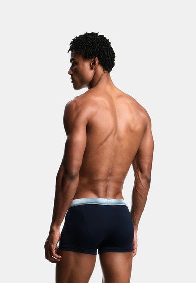 Muskulöser Mann mit dunkler Haut und lockigem Haar, der schwarze Boxershorts mit grauem Bund trägt und mit dem Gesicht nach links auf einem schlichten weißen Hintergrund steht.