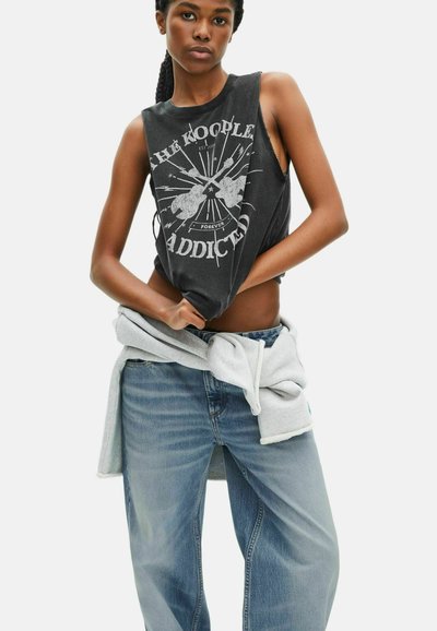 Zwarte mouwloze graphic tanktop met versleten randen, featuring witte tekst en ontwerpen. Gepaarde met lichtblauwe, loszittende denim jeans en een grijze hoodie.