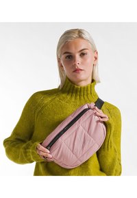 Geantă crossbody roz matlasată cu închidere cu fermoar, având un detaliu cu logo, purtată pe un pulover verde cu textură.