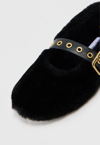 Sorte, fuzzy ballerinaer med rund tå, en udskæring på toppen og en sort læderrem med guldøjer og spænde. Stilen er både elegant og komfortabel.