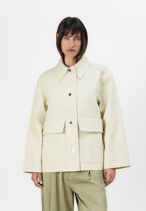 Beige overdimensjonert jakke med krage, to frontlommer og trykknapp-lukking. Glatt tekstur og strukturert design med synlige sømmer.
