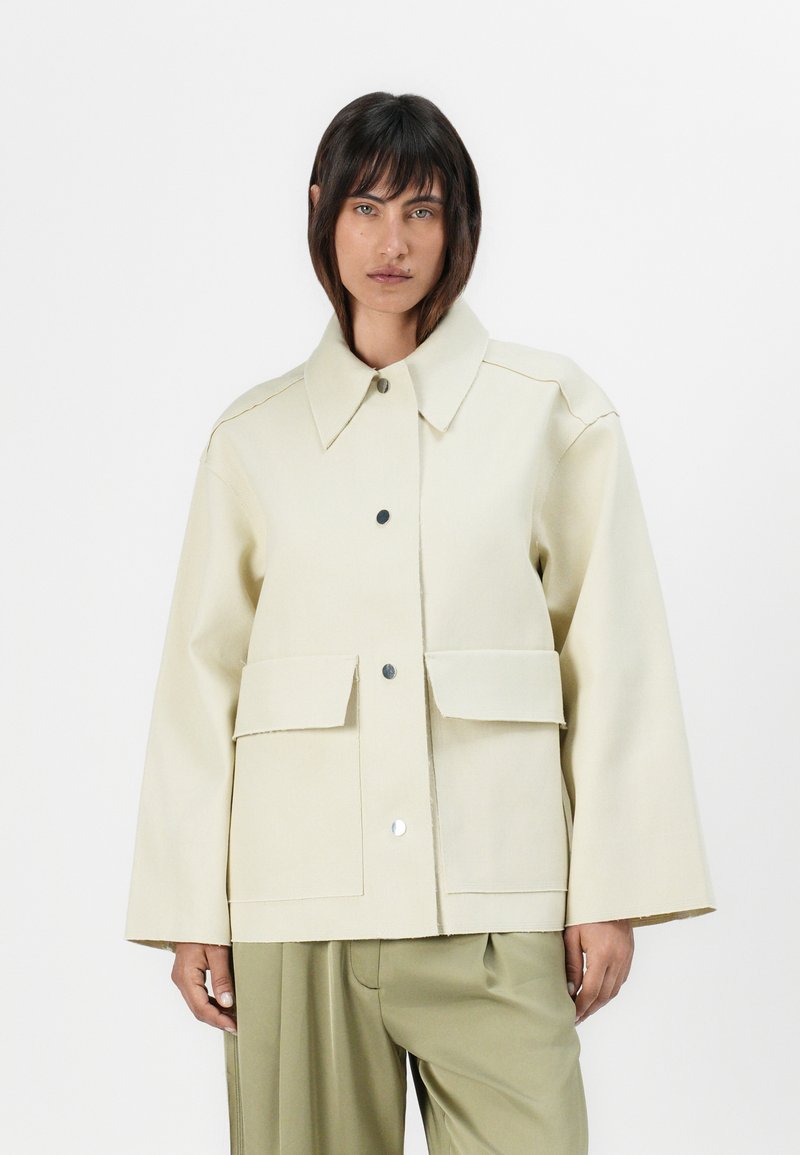 Beige oversized jacket with a collar, two front pockets, and snap-button closure. Smooth texture and structured design with visible seams.
