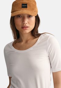 Vitt kortärmat t-shirt i mjukt tyg. Brun kordfläskaps med en svart "GANT"-etikett, böjd skärm och vertikalt ribbat mönster.