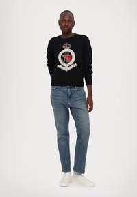 Zwarte gebreide trui met een logo ontwerp in rood, wit en grijs, gecombineerd met blauwe denimbroek en witte sneakers.