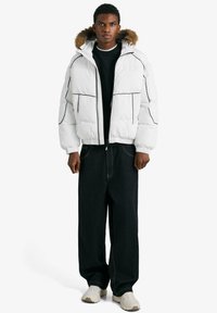 Biela prešívaná bunda s čiernymi obrysmi a kapucňou lemovanou kožušinou, v kombinácii s voľnými tmavými džínsami a béžovými teniskami. Jednoduchý dizajn, prešívaná textúra.