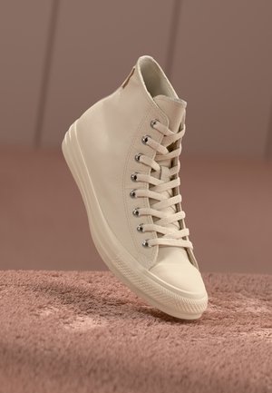 Baskets montantes beiges avec lacets, œillets métalliques et semelle en caoutchouc sur une surface texturée marron douce.