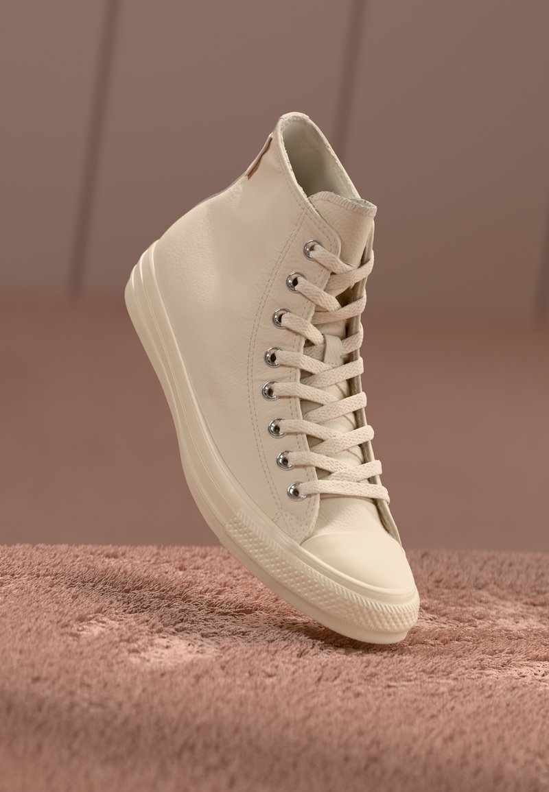 Baskets montantes beiges avec lacets, œillets métalliques et semelle en caoutchouc sur une surface texturée marron douce.