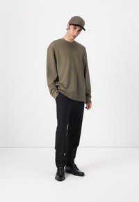 Pulover verde măsliniu cu mânecă lungă, croială lejeră, purtat cu pantaloni negri și pantofi din piele negri. O pălărie în carouri completează ținuta.