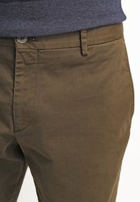 Hnědé bavlněné chinos s hladkou texturou, s klasickým designem pěti kapes a knoflíkovým zapínáním v pase.