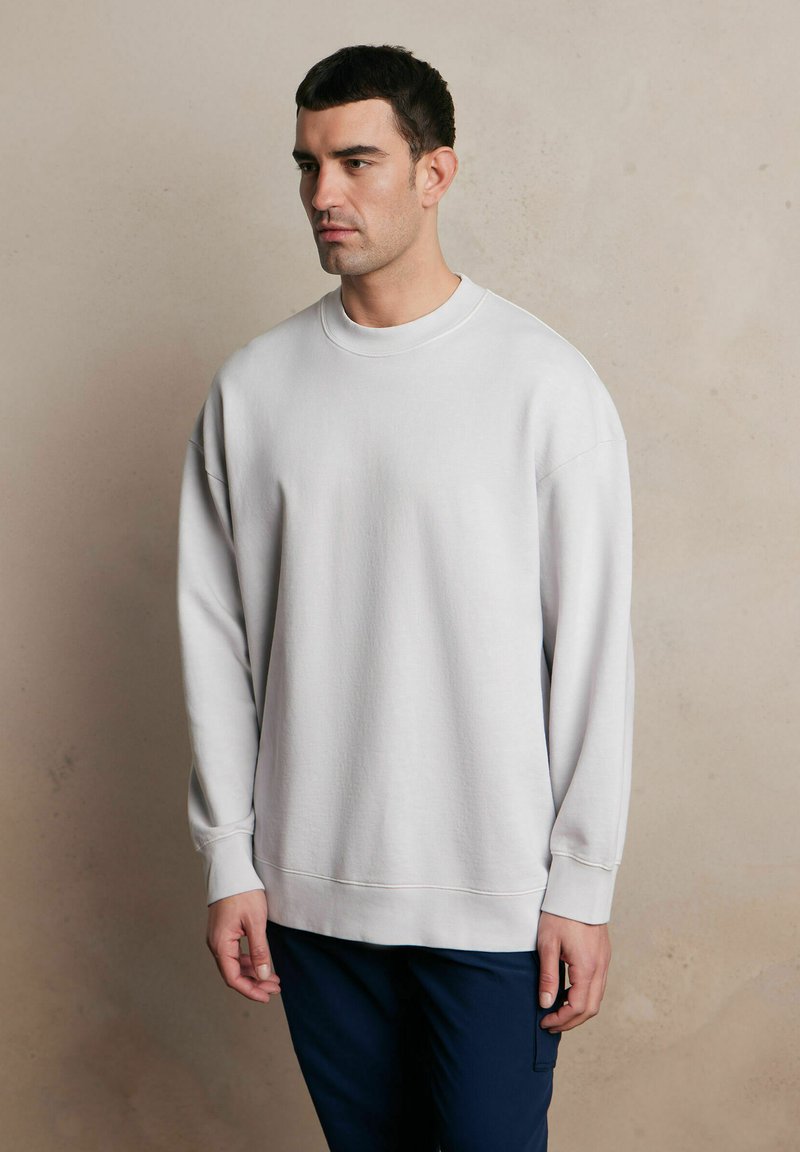 Jasnoszara bluza z okrągłym dekoltem, długimi rękawami i ściągaczami. Miękka tekstura i luźny krój, połączona z granatowymi spodniami.