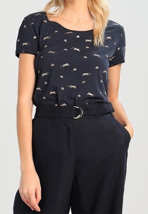 Vrouw die een zwart t-shirt draagt met kleine beige insectenpatronen, ingestopt in een zwarte broek met hoge taille en een dubbele ringriem.