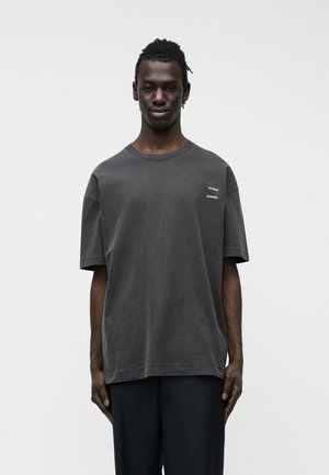 Junger Mann mit dunkler Haut, trägt ein lockeres dunkelgraues T-Shirt mit kleinem weißem Text auf der Brust und schwarze Hose, steht vor einem schlichten Hintergrund.