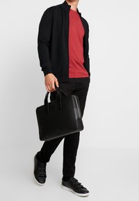 Czarna torebka na zamek błyskawiczny z dwoma rączkami, o teksturowanej powierzchni, zestawiona z czerwoną koszulką i czarnym kardiganem oraz spodniami, do tego czarne trampki na płaskiej podeszwie.