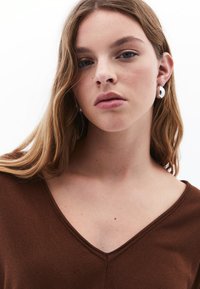 Braunes V-Ausschnitt-Oberteil aus weichem Stoff, das eng anliegt. Die Haare sind wellig und das Model trägt große silberne Ohrringe.