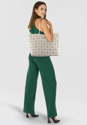 Capiente borsa a tracolla beige con un motivo a fantasia, dotata di doppie maniglie e chiusura a fibbia, abbinata a una tuta verde e tacchi neri.