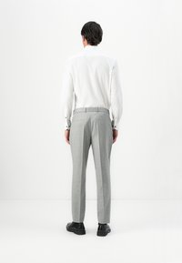 Man standing facing away wearing a white dress shirt, light grey tailored trousers, and black dress shoes against a plain background.