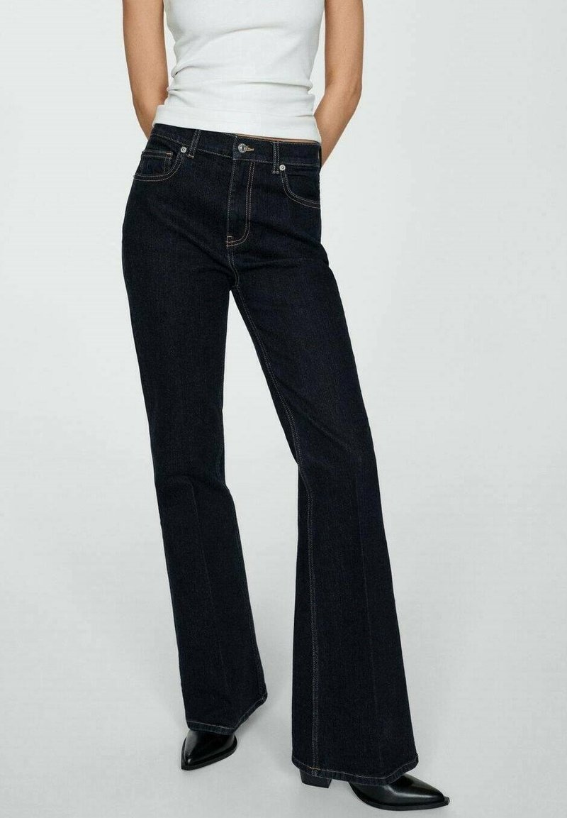 Vaqueros acampanados de color azul oscuro, confeccionados en denim, con bolsillos cosidos y cierre de botón. Combinados con una camiseta sin mangas blanca ajustada y tacones negros.