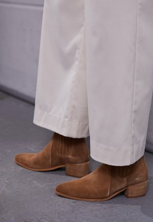 Brązowe zamszowe botki za kostkę z teksturowanym elastycznym panelem i prążkowanym detalem, zestawione z białymi spodniami o szerokich nogawkach.
