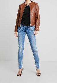 Brun läderjacka med rund krage, svart t-shirt, ljusblå skinny jeans och svarta snörsandaler, visad mot en neutral bakgrund.