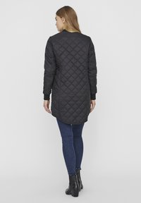 Veste longue matelassée noire avec un ourlet arrondi et des poignets côtelés, portée sur un jean foncé et des bottines. Texture lisse, détails minimalistes.