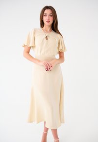 Midilanges, beiges Kleid aus weichem Stoff mit flatternden Ärmeln und einem Schlüsselloch-Detail am Ausschnitt. Fließt sanft bis zum Saum.