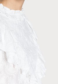 Vit blus med strukturerad tyg med blommönster, en volangkrage och intrikata detaljer, designad för en figurnära silhuett.