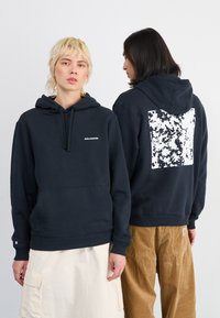 Tumesinine kapuutsiga sweatshirts koos ees pocketsiga. Rinnas on valge Salomoni logo ning seljal on tekstureeritud valge ja musta disain.