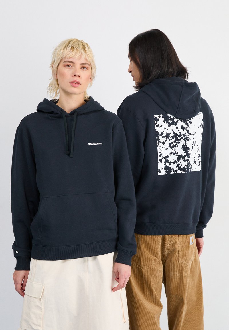 Tumesinine kapuutsiga sweatshirts koos ees pocketsiga. Rinnas on valge Salomoni logo ning seljal on tekstureeritud valge ja musta disain.