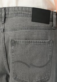 Acercamiento al bolsillo trasero de unos pantalones de mezclilla grises y el lazo del cinturón con una etiqueta de marca de cuero negro en la cinturilla, contra una camisa blanca.